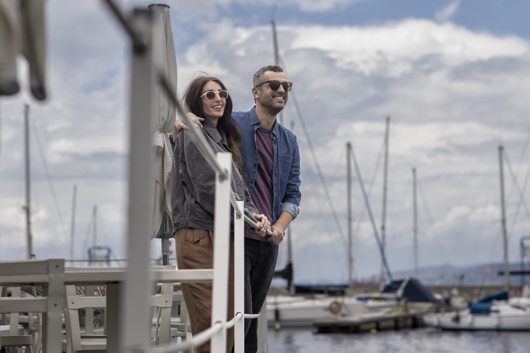 A couple is stading at the harbour with several sailing boats in the back and they are watching the boats leaving and coming into the harbour on a sunny day.