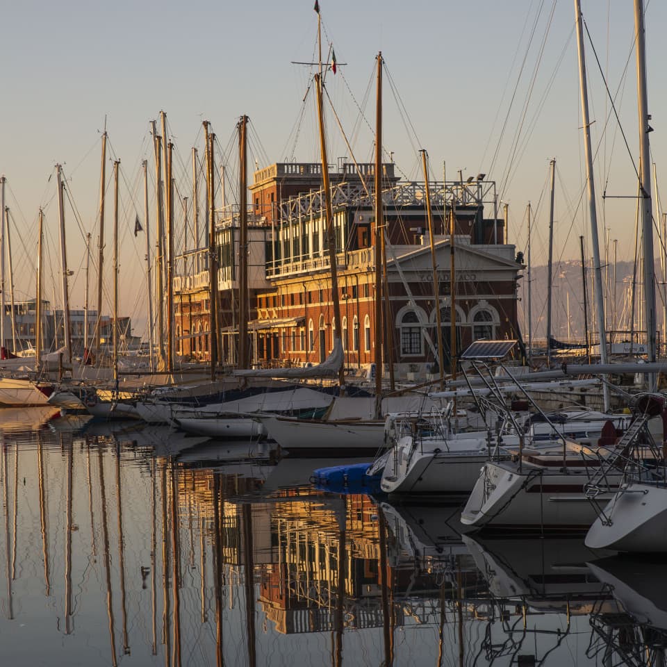 Blick auf die vielen Segelboote im Hafen von Triest bei Sonnenuntergang, mit einem Lagerhaus im Hintergrund.